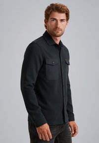 Črna srajca z dolgimi rokavi in gumbi, z teksturiranim zaključkom, dvema prsnikoma in ovratnikom, nosita čez temne kavbojke.