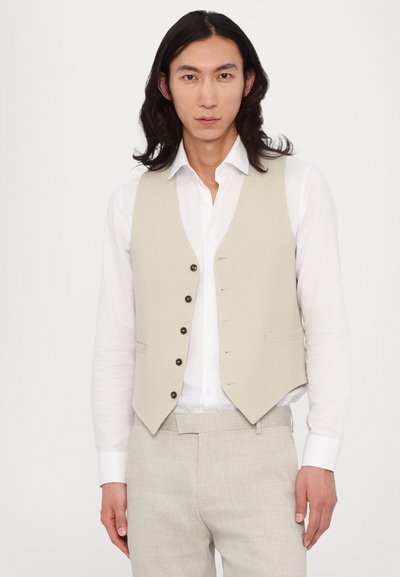 Hellbeige Leinenweste mit fünf schwarzen Knöpfen, zwei vorderen Taschen und slim fit. Getragen über einem weißen Hemd und hellbeigen Hosen.