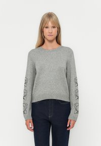 Grå strikkegenser med lange ermer, med svart blomsterbroderi på ermene, rund halsutskjæring og ribbestrikket kant. Kombinert med mørke jeans.