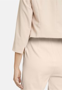 Lichtbeige cropped jack met een gladde textuur, zijslitsen en driekwartmouwen, gecombineerd met taps toelopende broeken in dezelfde kleur.