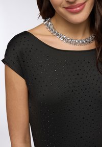 Top nero con scollatura ampia, decorato con borchie di diverse dimensioni in nero e argento, abbinato a una collana di cristalli trasparenti.