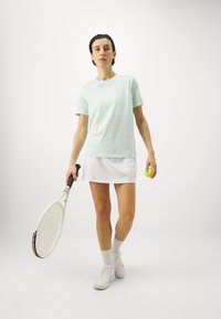 T-shirt em algodão verde menta, de mangas curtas, combinado com uma saia branca. A pessoa segura uma raquete de ténis e uma bola de ténis amarela.