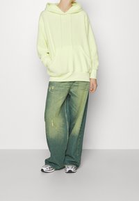 Sudadera verde lima con bolsillo frontal y cordones, combinada con vaqueros azules sueltos y descoloridos que presentan detalles desgastados, y zapatillas deportivas.