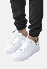 Bílé tenisky Nike Air Force 1 s perforovanou špičkou, plochými tkaničkami a texturovanou gumovou podešví, kombinované s černými jogger kalhotami.