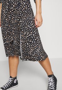 Schwarzes Kleid mit einem gepunkteten Muster in Weiß und Beige, mit einem leichten Schlitz am Saum. Kombiniert mit schwarzen High-Top-Sneakern.