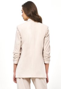 Kobieta z długimi brązowymi włosami, ubrana w jasnobeżową marynarkę z marszczonymi rękawami oraz pasującymi spodniami, stojąca tyłem na białym tle.