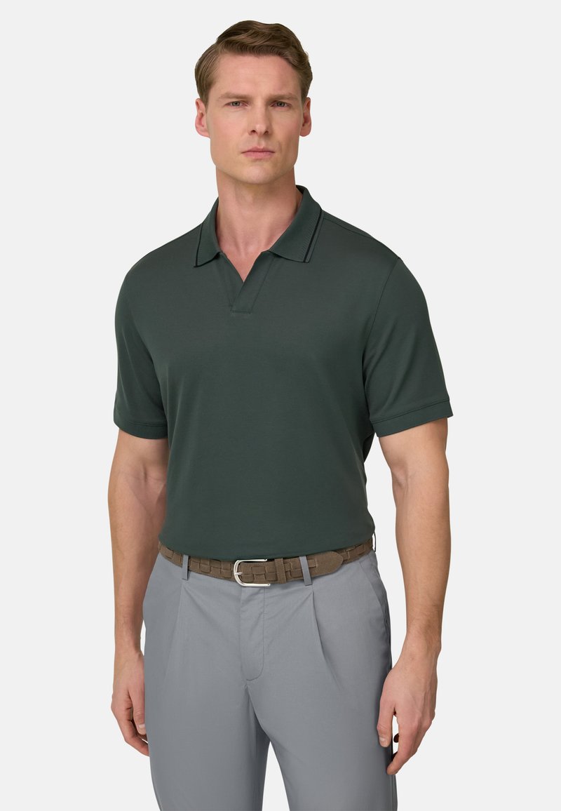 Man draagt een donkergroen poloshirt met korte mouwen, ingestopt in lichtgrijze broek met een bruine riem, staand tegen een effen achtergrond.