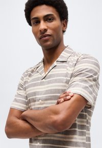 Άνδρας με σταυρωμένα χέρια, φορώντας πουκάμισο με κοντά μανίκια και οριζόντιες ρίγες σε μπεζ και άσπρο, κοιτάζει με αυτοπεποίθηση την κάμερα.