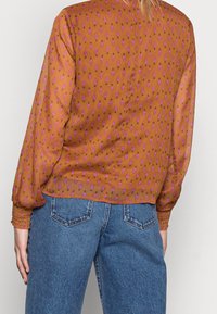 Braune Bluse mit einem floralen Muster in Rosa, mit langen Ärmeln und lockerer Passform. Getragen mit hellblauen hoch taillierten Jeans.