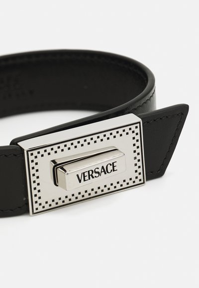 Melna ādas aproce ar taisnstūrveida sudraba sprādzi, kurai ir rakstainas akcenti un iegravēts "VERSACE" logos.