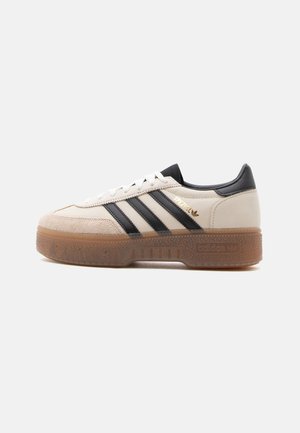 Zapatilla blanca y beige con rayas negras, lengüeta trasera negra, etiqueta "Spezial" en dorado y suela marrón translúcida y gruesa.