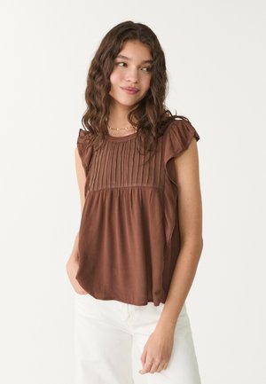 Brown pleated top with short ruffled sleeves, lightweight fabric, and flared hem, worn with white pants, featuring a round neckline.