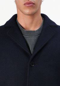 Cappotto di lana navy con collo a revers, chiusura a bottone singolo e un maglione grigio a girocollo sotto. Texture liscia e vestibilità sartoriale.