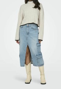 Femme portant un pull crème avec un bord noir, une jupe mi-longue en denim bleu clair avec des poches cargo et une fente à l'avant, ainsi que des bottes de cowboy crème.
