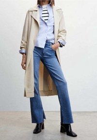 Beiger Trenchcoat über einem hellblauen Hemd mit schwarzen Streifen, kombiniert mit blauen Schlagjeans. Schwarze Stiefeletten vervollständigen das Outfit.