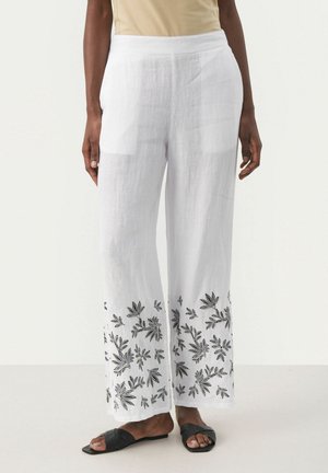 Witte broek met wijde pijpen en zwarte bloemembroderie langs de zoom, gedragen met een beige top en zwarte sandalen met open teen.