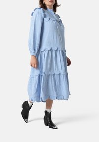 Vestido de cuadros azul y blanco con cuello y mangas voladas, diseño en capas, dobladillo festoneado, combinado con botas de tobillo negras con puntas metálicas.