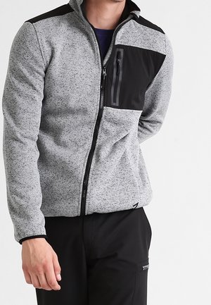 Mand iført en grå fleecejakke med sort brystlomme og sorte bukser, stående mod en hvid baggrund.