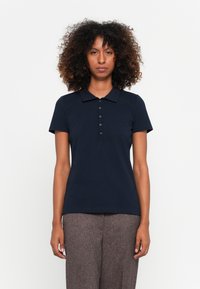 Marineblaues Polohemd mit kurzen Ärmeln, Knopfverschluss und Kragen. Trägt man mit braunen strukturierten, hochgeschnittenen Hosen.