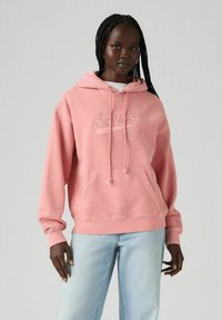 Sudadera rosa con un bolsillo delantero, capucha con cordón y logo en relieve de "Levi's". Combinada con jeans de mezclilla azul claro. Textura suave.