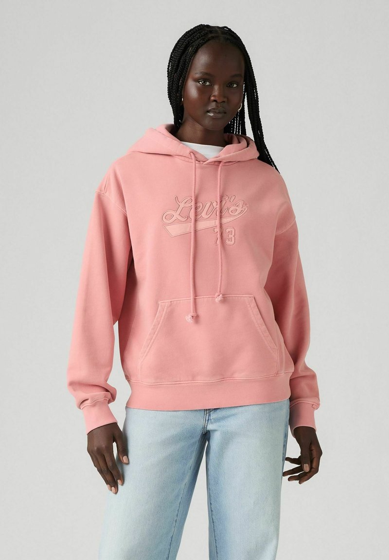 Sudadera rosa con un bolsillo delantero, capucha con cordón y logo en relieve de "Levi's". Combinada con jeans de mezclilla azul claro. Textura suave.