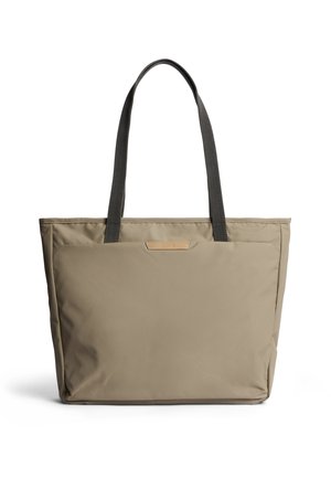 Borsa a mano beige con spalline nere, tasca frontale rettangolare e design minimalista su sfondo bianco.
