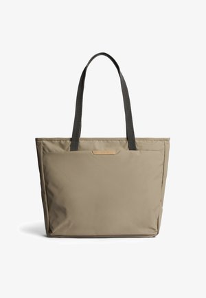 Borsa a mano beige con spalline nere, tasca frontale rettangolare e design minimalista su sfondo bianco.