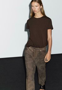 Woman with long hair walking, wearing a dark brown short-sleeve shirt, brown suede pants, a silver ring, and black sandals.