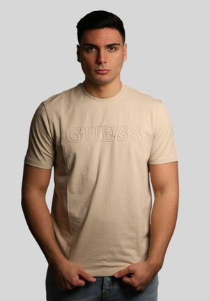 Giovane uomo con capelli corti e scuri che indossa una maglietta beige GUESS e jeans blu, in piedi davanti a uno sfondo grigio chiaro uniforme.