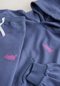 Sudadera y pantalones de chándal azul marino, confeccionados en tela suave. Presentan puños acanalados y un logo bordado en rosa en ambas prendas.
