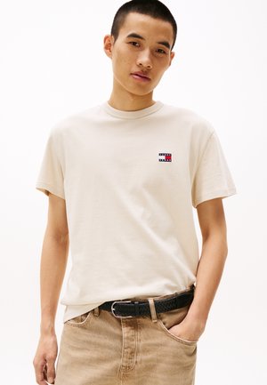 Camiseta de algodón beige con cuello redondo y mangas cortas. Presenta un pequeño logotipo rectangular en rojo, blanco y azul en el pecho.