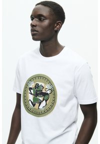 White short-sleeve t-shirt featuring a circular graphic design with black, green, and tan elements depicting figures and patterns.