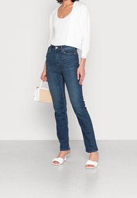 Jeansy z wysokim stanem w niebieskim kolorze, z delikatnym streczem, zestawione z białym, krótkim kardiganem i białymi sandałami na blokowym obcasie.