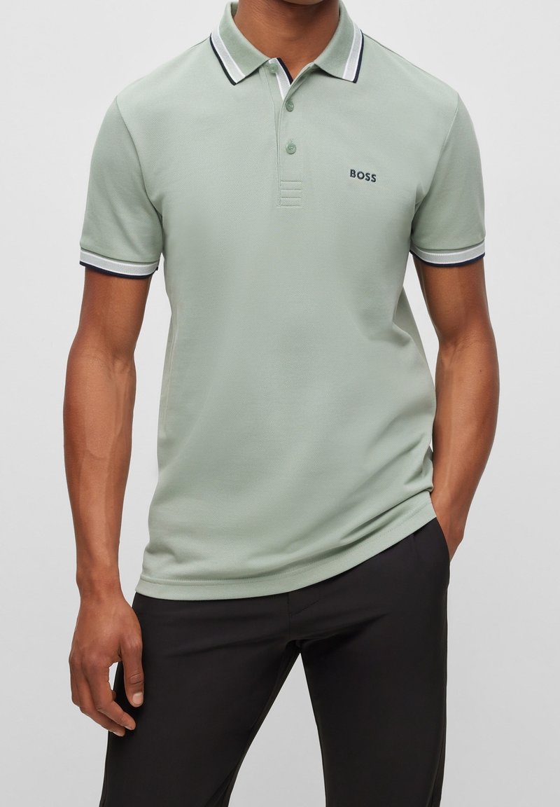 Homme portant un polo à manches courtes vert clair avec bordures noires et blanches et un pantalon noir, debout avec une main dans la poche.