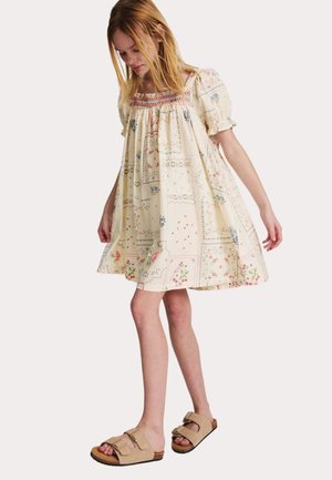 Młoda osoba ubrana w luźną kremową sukienkę z kwiatowymi wzorami i beżowe sandały na podwójnych paskach, stojąca z lekkim spuszczonym wzrokiem.