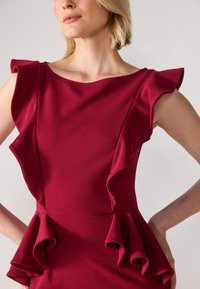 Burgunderrød kjole laget av teksturert stoff, med volanger på ermene og en lagdelt peplum i midjen, med en klassisk båtnek.