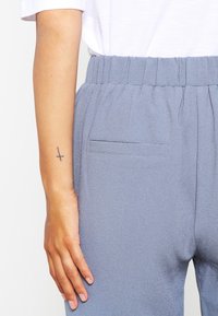 Ljust blå, texturerade byxor med elastisk midja och bakficka; hand som vilar på midjan; synlig tatuering på handleden.