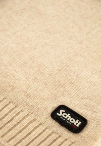 Tissu tricoté beige avec des bords côtes et une étiquette rectangulaire noire de la marque Schott NYC cousue sur le coin inférieur droit.