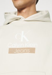 Felpa beige realizzata in tessuto morbido, con un ampio logo bianco riportante "Calvin Klein" e "Jeans" in lettere maiuscole marroni.