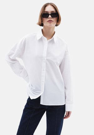 Camicia bianca con bottoni realizzata in cotone fresco, con colletto classico e vestibilità rilassata, abbinata a jeans blu scuro e occhiali da sole neri rettangolari.