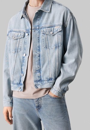 Veste en jean bleu clair et jean portés sur un T-shirt beige, avec une main dans la poche et le logo Calvin Klein visible sur la poche de la veste.