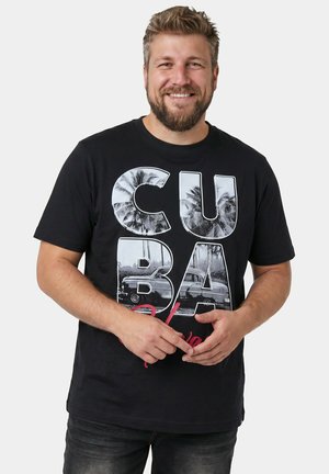 Lächelnder bärtiger Mann in schwarzem "CUBA" T-Shirt mit Palmen und klassischen Autos, stehend vor hellem, einfarbigem Hintergrund, Hände locker verschränkt.