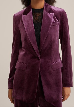 Modèle portant un blazer en velours couleur prune sur un haut noir en dentelle, avec un pantalon assorti. Le blazer est doté de larges revers et de poches à rabat.