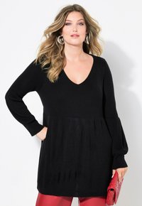 Tunic nera a maglia con scollo a V, maniche lunghe a sbuffo e orlo fluente. Abbinata a pantaloni rossi e una borsa rossa con texture.