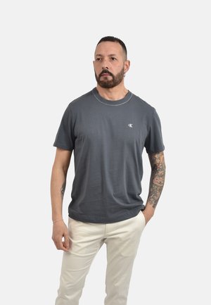 Uomo con barba e braccio destro tatuato che indossa una t-shirt Calvin Klein grigio scuro e pantaloni beige chiaro, in piedi con una mano in tasca.