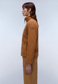 Fleecejacke in Hellbraun mit hohem Kragen, Reißverschlusstaschen und strukturiertem Stoff. Mit dezentem Branding auf der Brust und am Saum.
