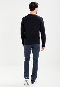 Långärmad svart bomullsskjorta kombinerad med marinblå byxor och vita sneakers, sedd bakifrån, vilket framhäver den enkla, avslappnade designen.