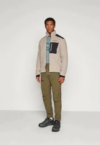 Casaco de fleece bege claro com detalhes em azul-marinho, com bolso de peito com fecho, usado sobre uma camisa azul e calças verde-oliva, combinado com sapatos pretos.