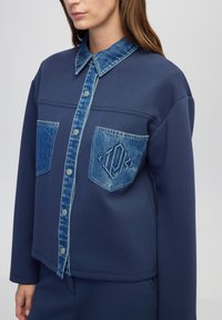 Blaues Sweatshirt-Jacket mit Denim-Akzenten, ausgestattet mit einer durchgehenden Knopfleiste, zwei Brusttaschen und einem strukturierten Kragen. Weicher Stoff.