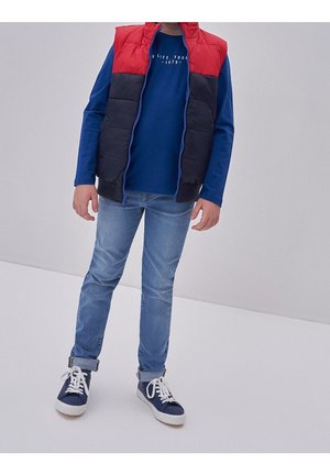 Dítě oblečené v modrém tričku s dlouhým rukávem, červeno-černé bezrukávé péřové vestě, světlých modrých džínách s ohrnutými lemy a modrých teniskách s bílými tkaničkami.